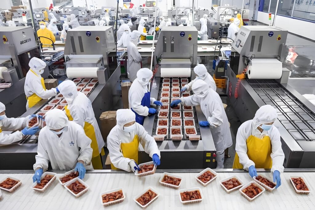Workers in full hygienic protective gear (white coveralls, masks, hairnets, and blue gloves) processing and packaging ready-to-eat meat products on a semi-automated production line in a modern food factory.