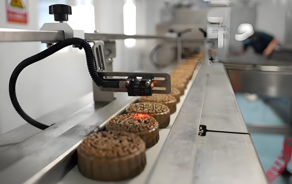 Mooncakes moving along an automated food packaging production line, with a laser inspection or marking sensor positioned above for quality control in a hygienic food factory.