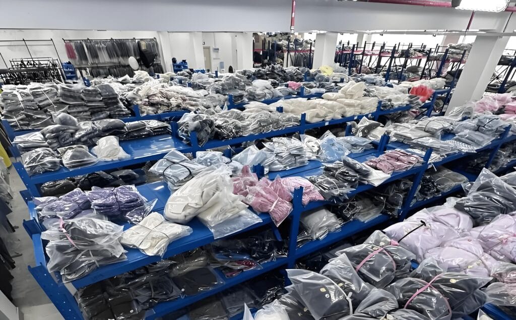 Hand-folded apparel inventory neatly arranged on blue shelves in a warehouse, showing organized finished garments with high-quality manual folding, ready for packaging and shipping
